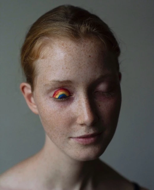 Close-up portrait of a red-haired adult female with freckles, wearing rainbow colored eyeshadow on one closed eye, against a grey background.