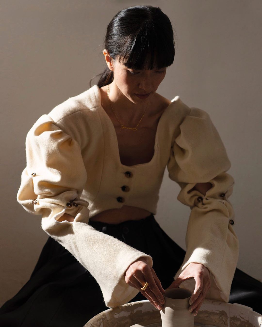 An Asian woman in a cream-colored puffy-sleeved top is seated at a pottery wheel shaping a clay pot in a naturally lit room.