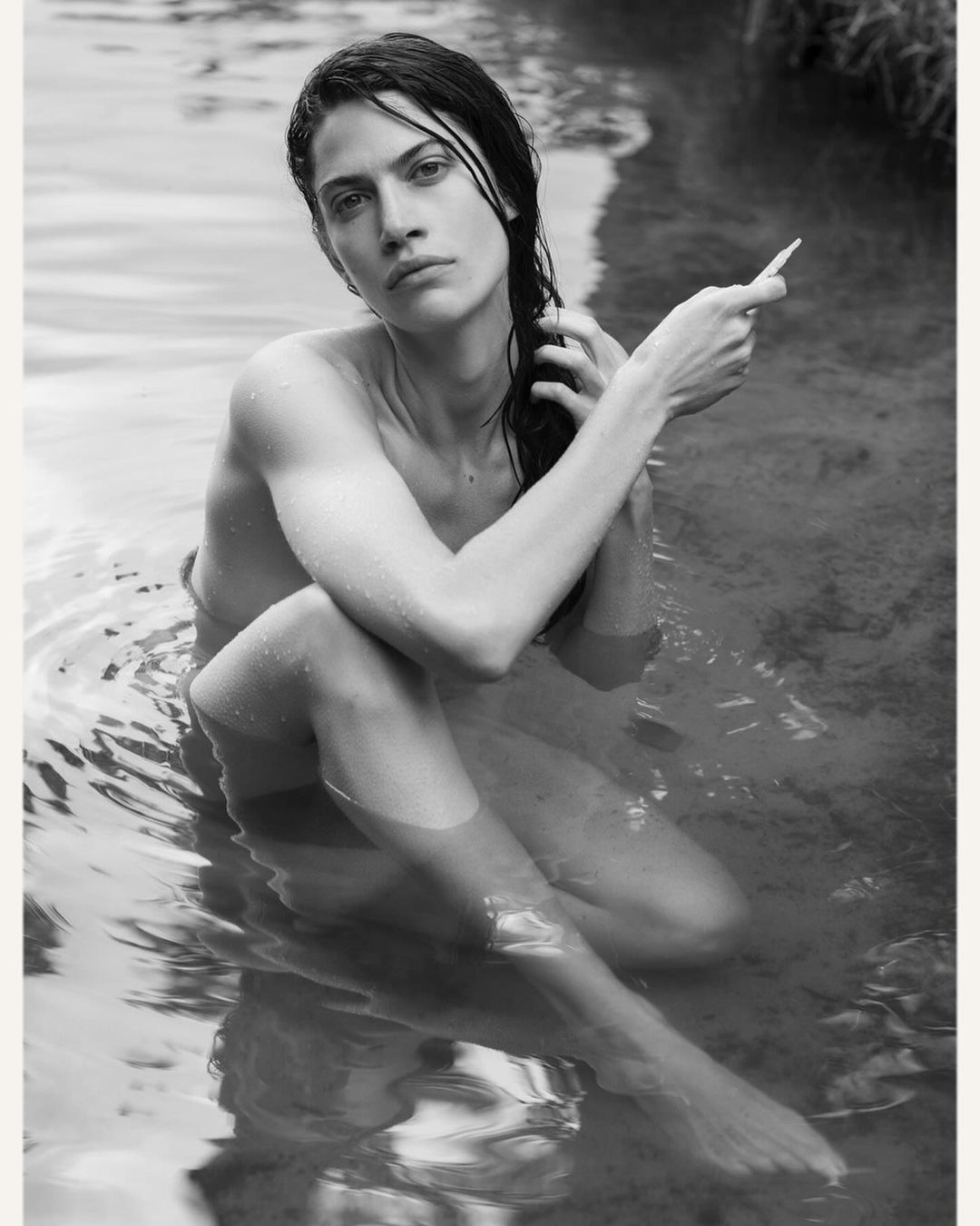 Black and white portrait of a fair-skinned adult woman sitting in water, her hand moving through her dark, wet hair.