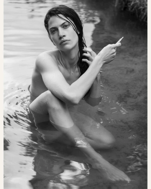 Black and white portrait of a fair-skinned adult woman sitting in water, her hand moving through her dark, wet hair.