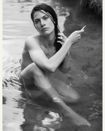 Black and white portrait of a fair-skinned adult woman sitting in water, her hand moving through her dark, wet hair.