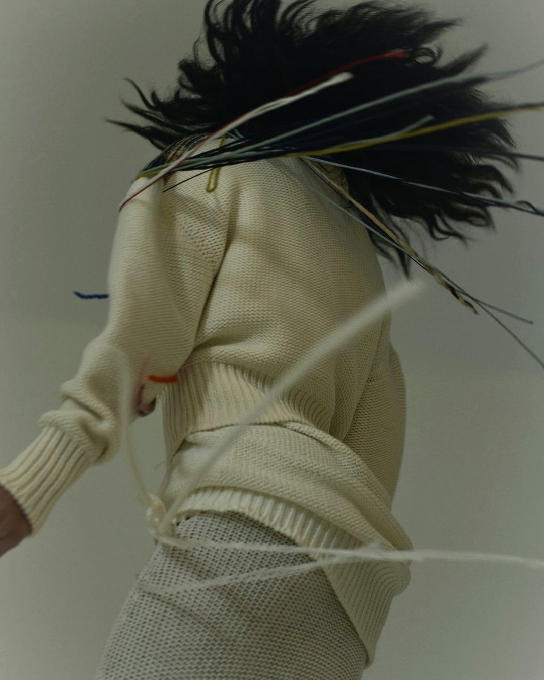 Woman in motion wearing a knit sweater and skirt, accented by loose threads and dynamic hair, captured in a studio setting.