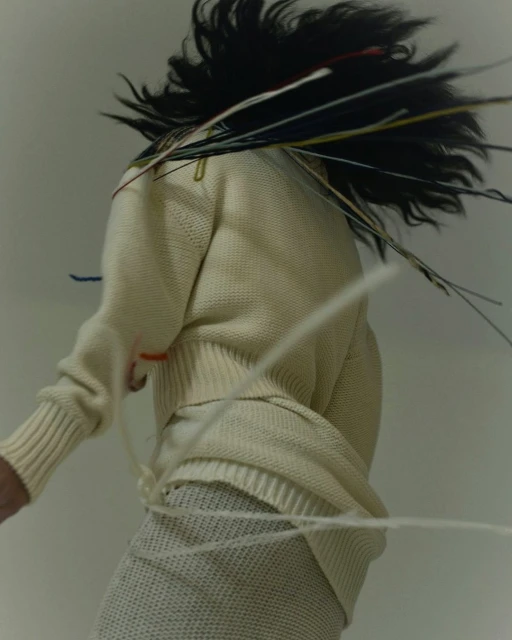 Woman in motion wearing a knit sweater and skirt, accented by loose threads and dynamic hair, captured in a studio setting.