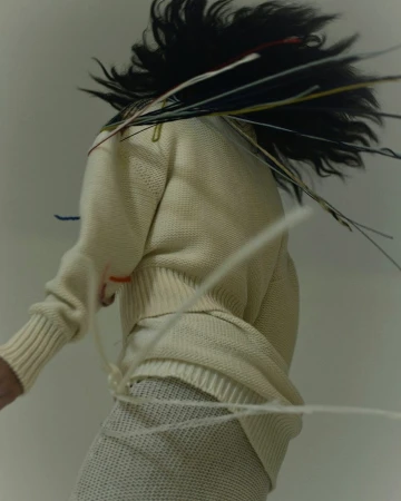Woman in motion wearing a knit sweater and skirt, accented by loose threads and dynamic hair, captured in a studio setting.