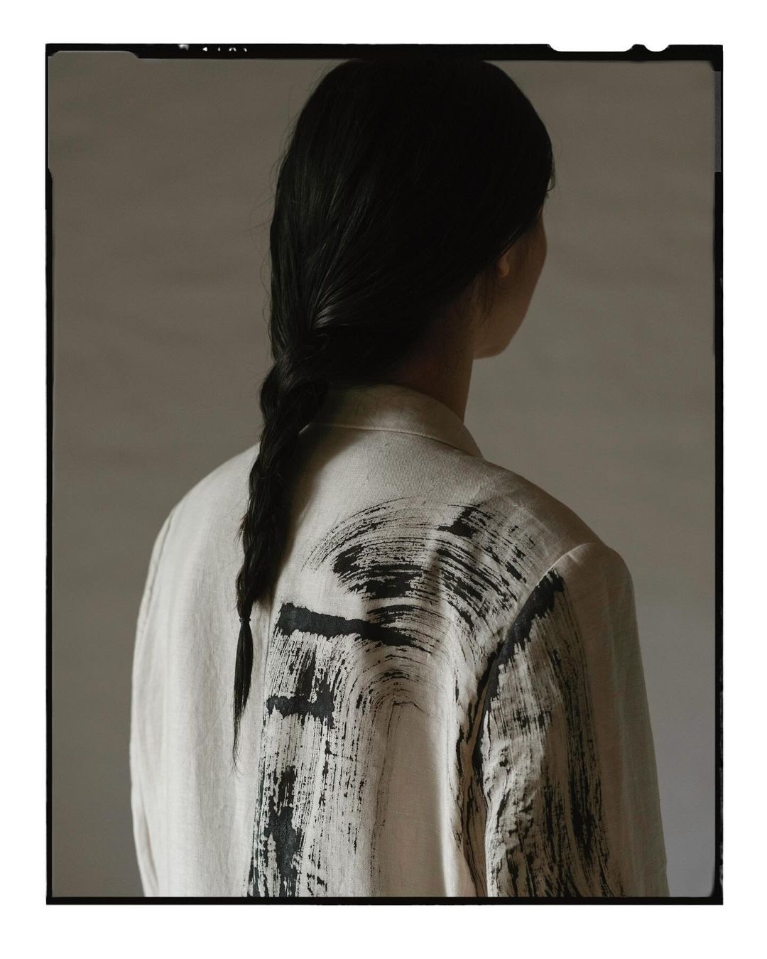 Back view of an adult female with a braid, wearing a light beige jacket featuring black brushstroke designs, set against a muted background.