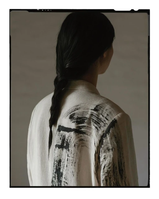 Back view of an adult female with a braid, wearing a light beige jacket featuring black brushstroke designs, set against a muted background.