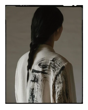 Back view of an adult female with a braid, wearing a light beige jacket featuring black brushstroke designs, set against a muted background.