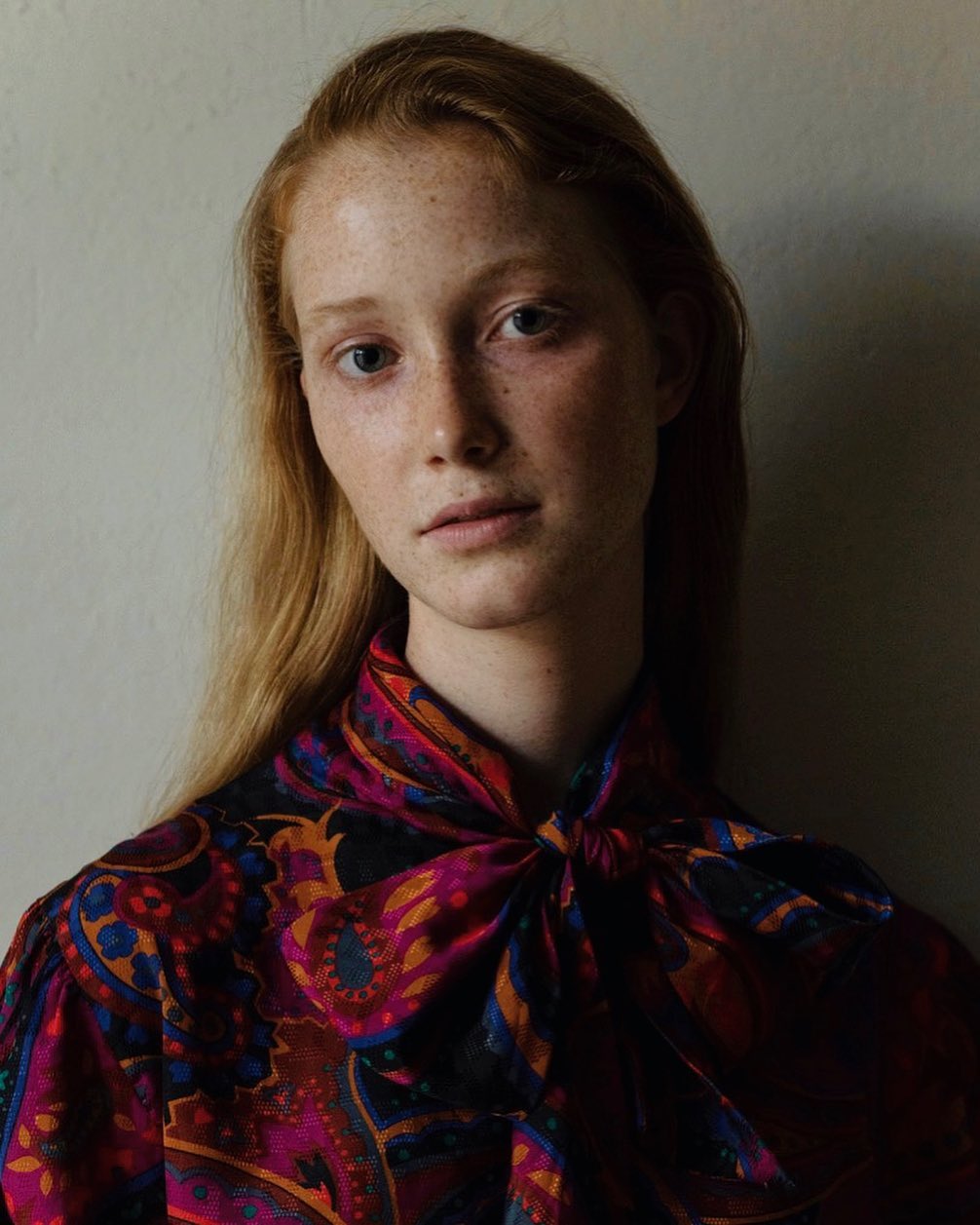 Portrait of a young woman with red hair and freckles wearing a patterned blouse, standing in front of a neutral wall with soft lighting.