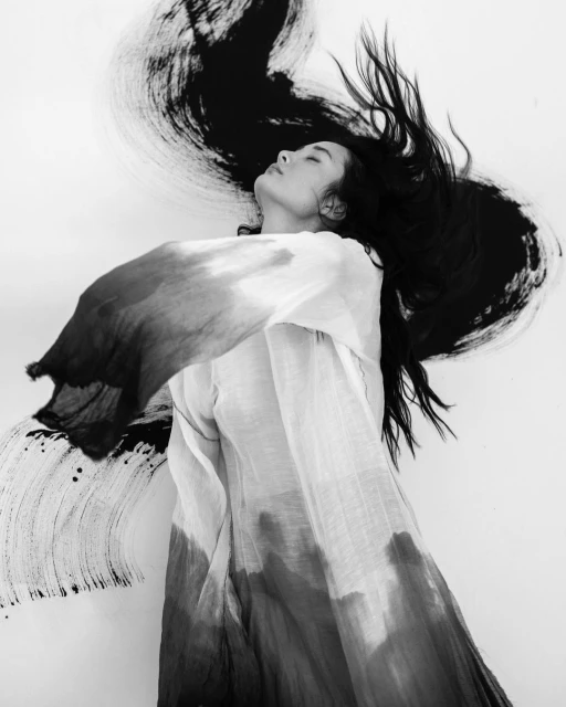 Black and white portrait of an Asian woman with long hair, wearing a draped white top, posing with a large artistic brushstroke in the background.