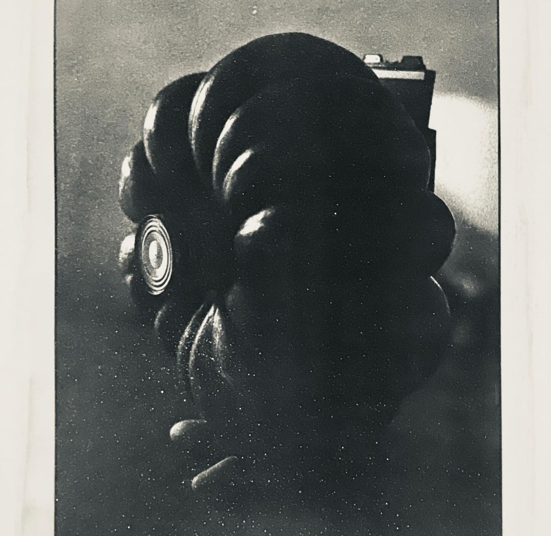 Close-up monochrome photograph of a Hasselblad camera with a Zeiss lens and a light source behind, set against a mottled backdrop.