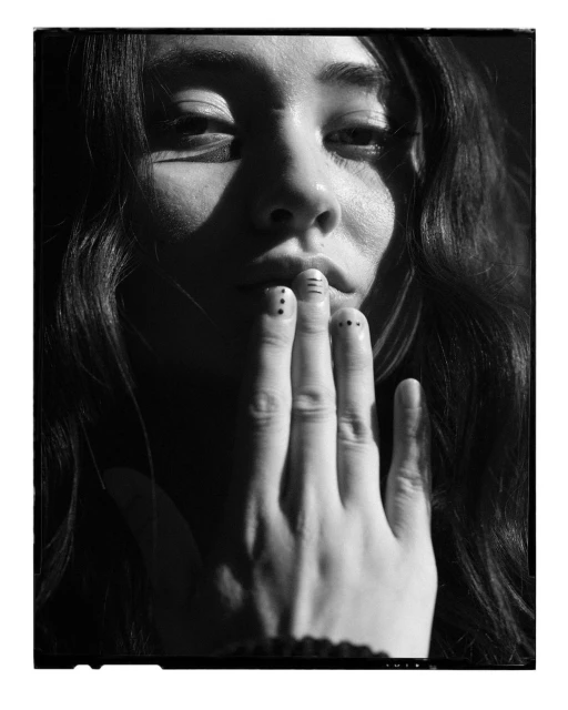 Portrait of a young adult with long dark hair partially obscuring her face with a hand displaying black dot nail art, in black and white.