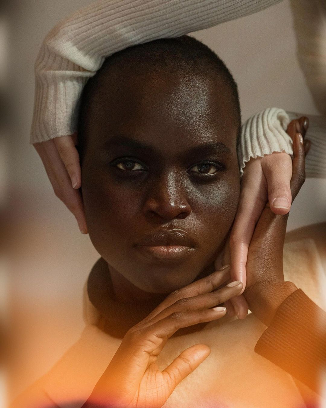 Close-up portrait of a woman with dark skin, fair hands framing her face, soft lighting, and a neutral expression.