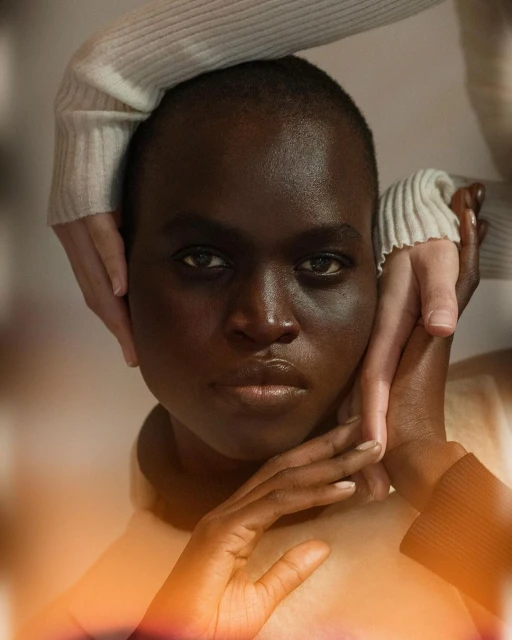 Close-up portrait of a woman with dark skin, fair hands framing her face, soft lighting, and a neutral expression.