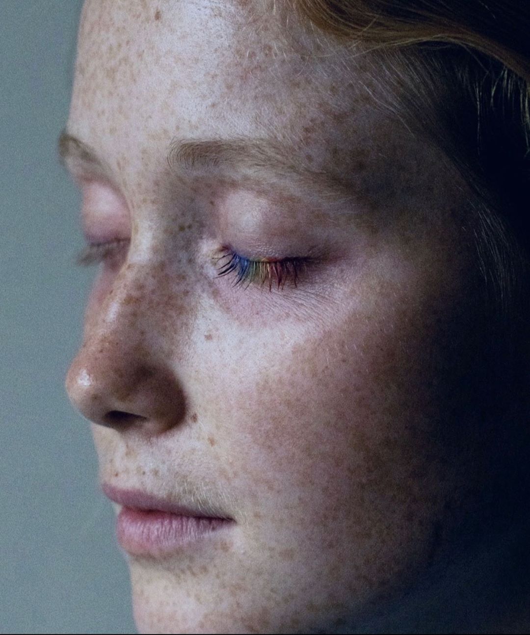 Close-up portrait of a fair-skinned woman with freckles and closed eyes with rainbow-colored eyelashes against a grey background.