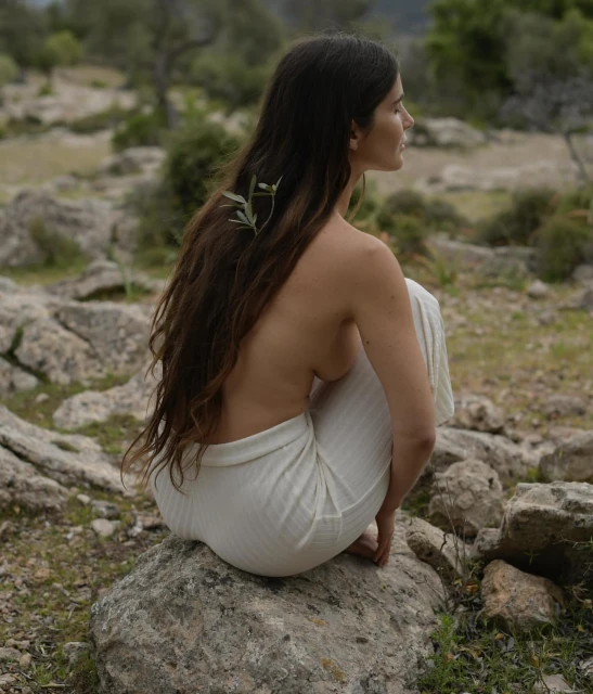 Brunette woman with bare upper body sitting on a rock in a forest environment, with an olive branch in her dark hair.
