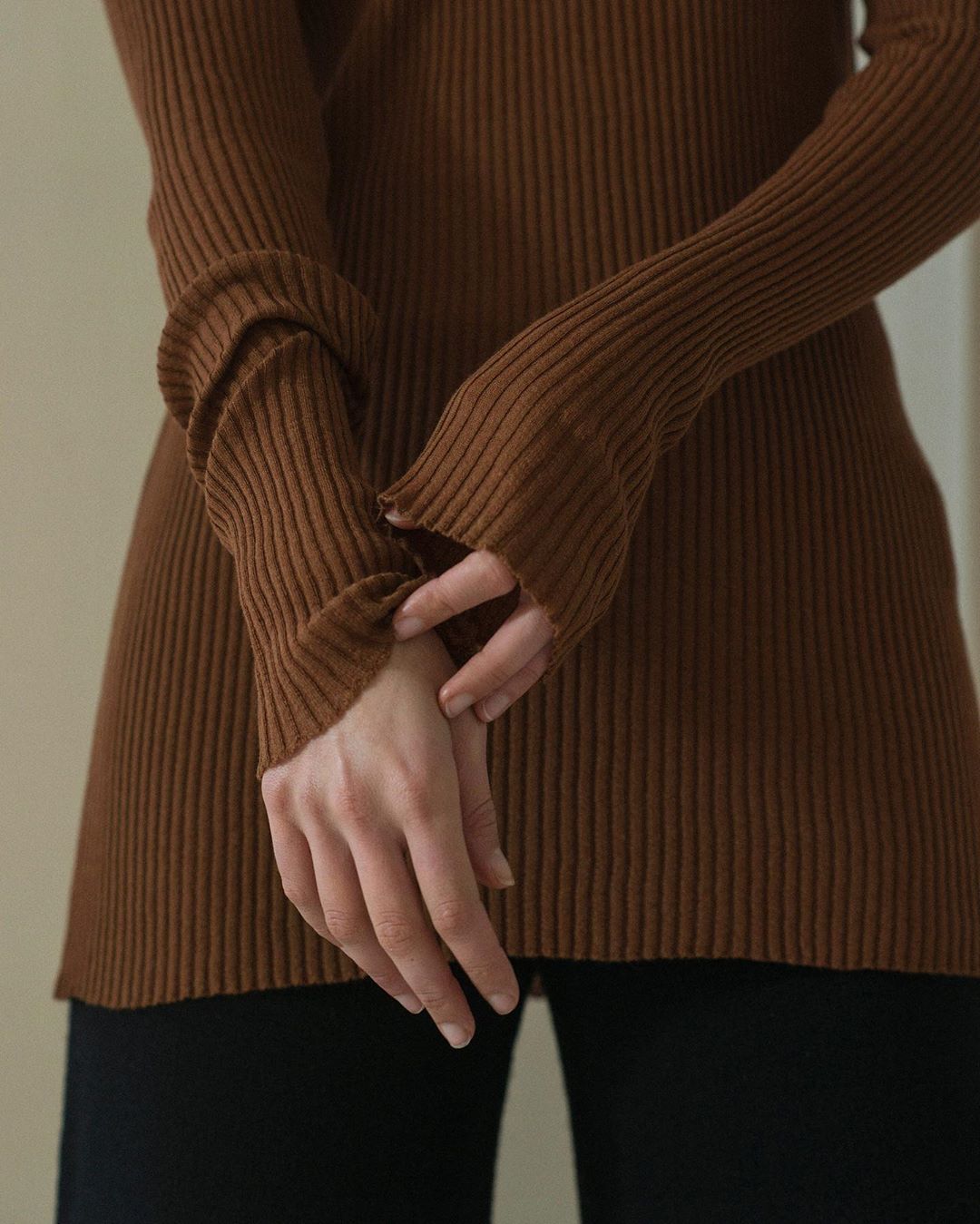 Close-up of a slender adult female wearing a fitted brown ribbed sweater and black pants in an indoor setting.