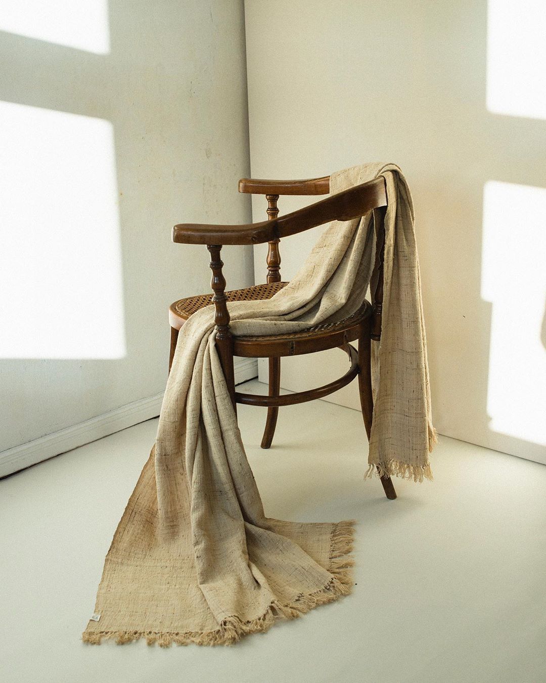 Wooden arm chair with a draped beige linen throw blanket. Neutral earthy tones, with natural light and shadow in an indoor setting.