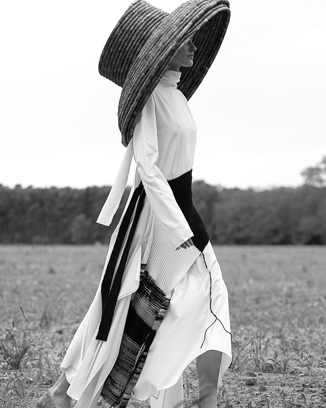 Monochrome outdoor shot of a fashion model in a wide-brimmed hat and layered white dress, walking through a grassy field under a bright sky.