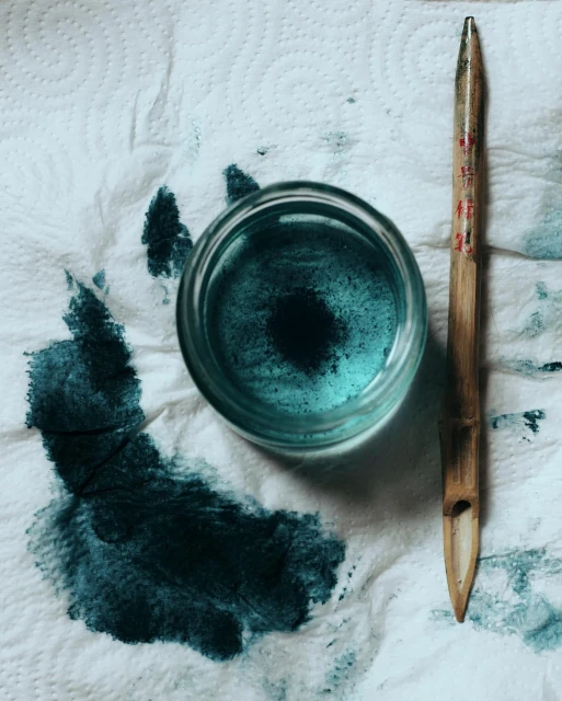 Close-up of calligraphy ink in a glass container with a bamboo calligraphy pen and ink spatter on a white paper towel.
