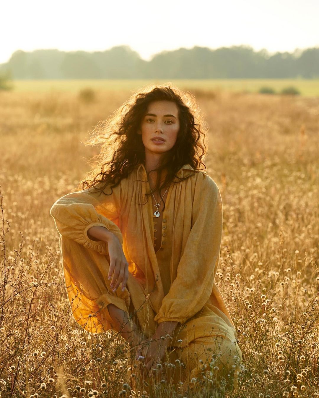 Woman with long curly hair in a golden dress sits in a sunlit field of tall grass, with a backdrop of trees and a clear sky.