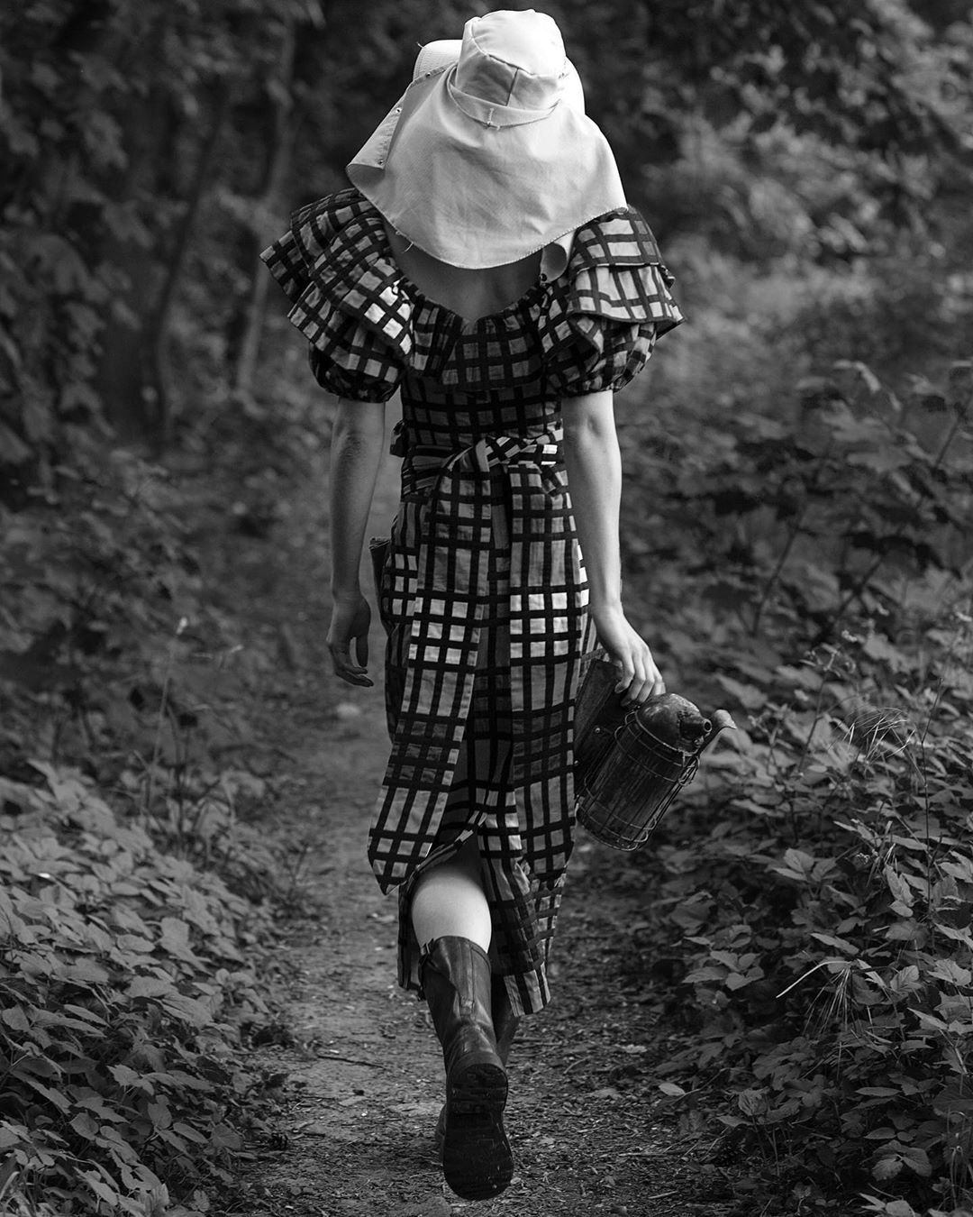 Back view of a person in a patterned midi dress, wide-brimmed hat, and boots on a dirt path through a lush, leafy forest, black and white.