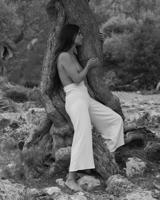 Monochrome portrait of a woman embracing a tree in nature, showcasing bare shoulders and wearing white pants in an outdoor setting.
