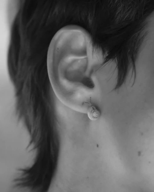 Close-up black and white shot of a person's ear with a silver earring. Focus on earlobe and simple earring design against dark hair.