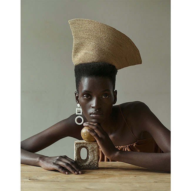 Portrait of a dark-skinned woman wearing a beige hat and earrings, resting her chin on a stone block with a gold ball on top.