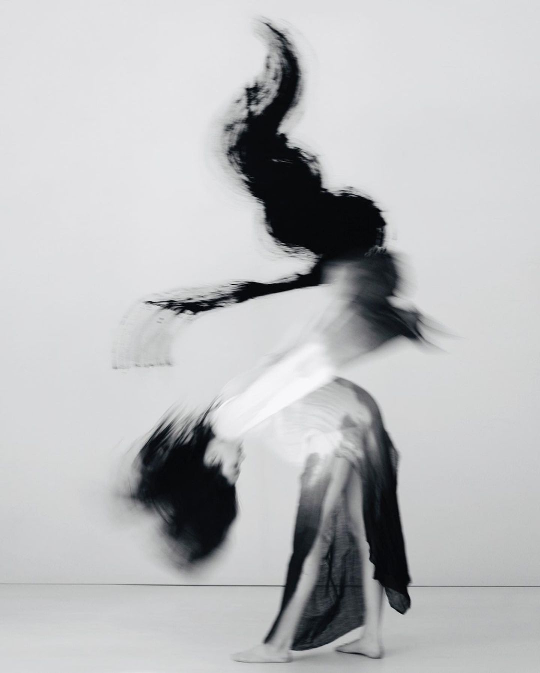 Abstract black and white long exposure shot of a person in motion wearing dark fabric against a white background, creating a blurred effect.