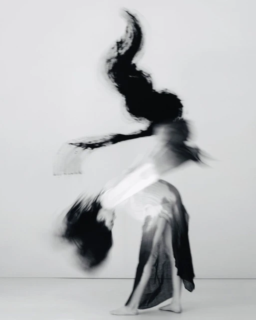 Abstract black and white long exposure shot of a person in motion wearing dark fabric against a white background, creating a blurred effect.