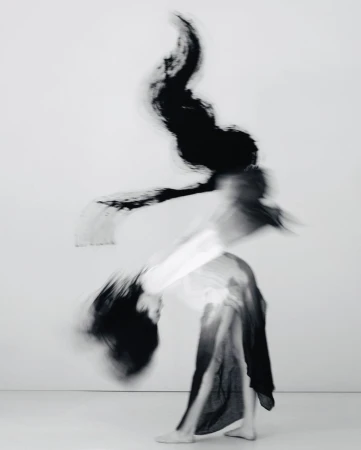 Abstract black and white long exposure shot of a person in motion wearing dark fabric against a white background, creating a blurred effect.