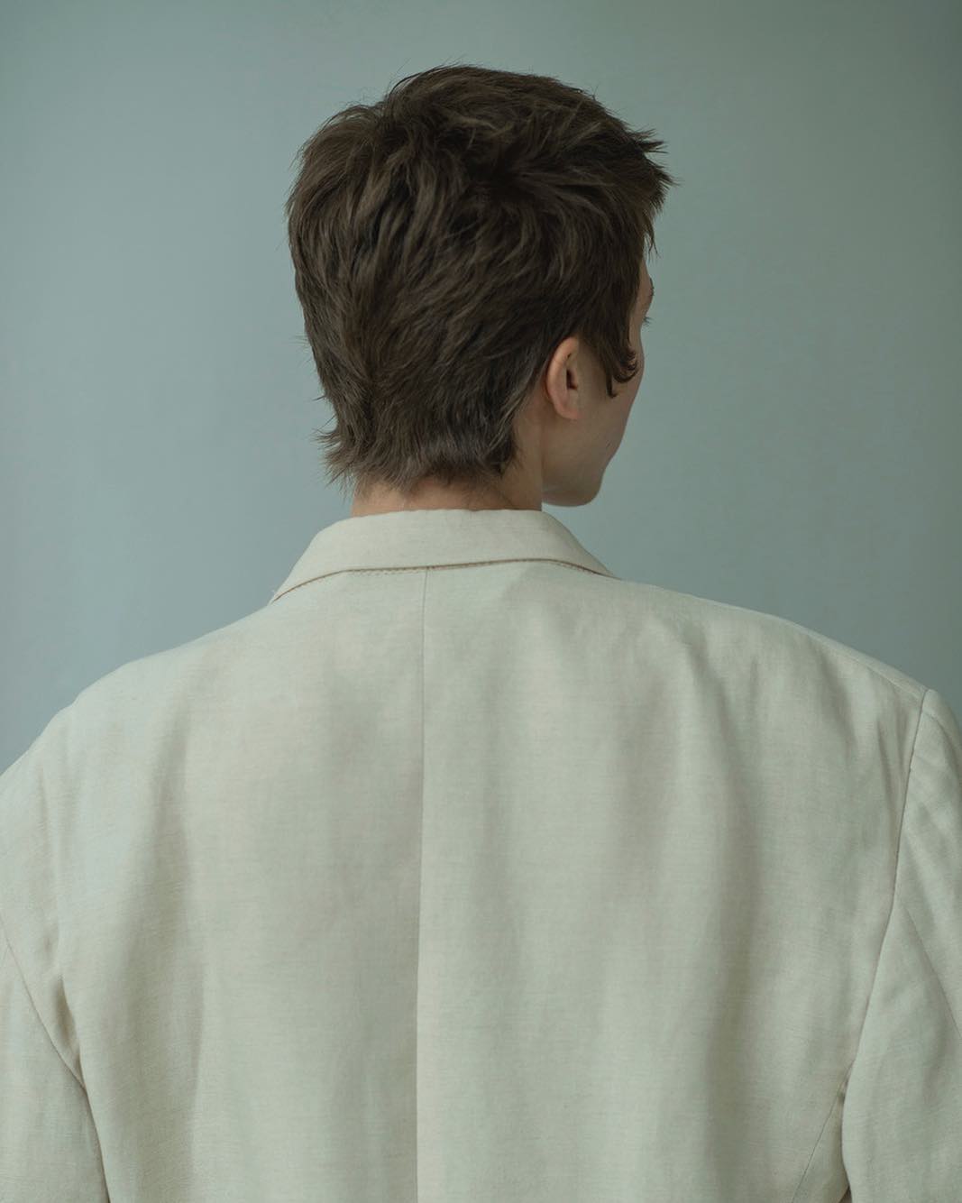 Back view of a person with short brown hair wearing a cream-colored blazer against a light grey background in a neutral indoor setting.