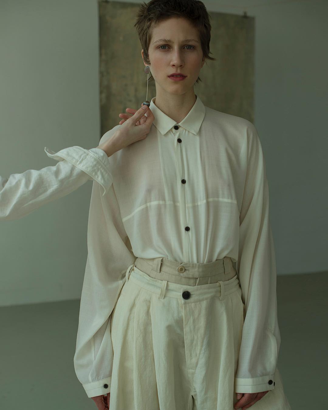 Fashion photo of a person with short hair wearing a white shirt and pants, standing indoors with a hand adjusting the collar.
