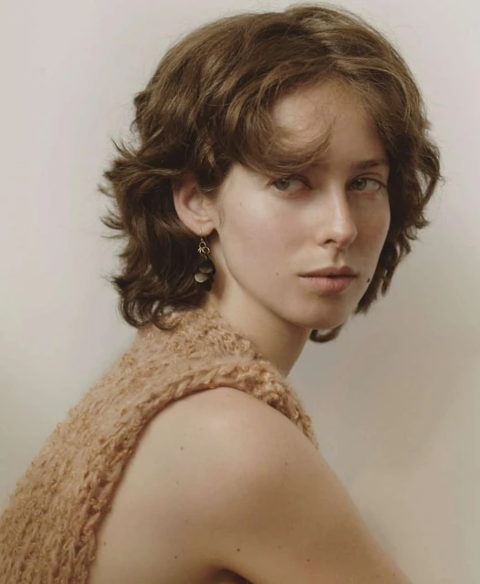 Portrait of a young adult female with a neutral expression, fair skin, and short brown hair, wearing an open-weave knit vest in a studio setting.