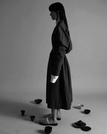 Monochrome portrait of a woman standing in profile wearing a long dark coat, surrounded by small dark bowls on the floor.