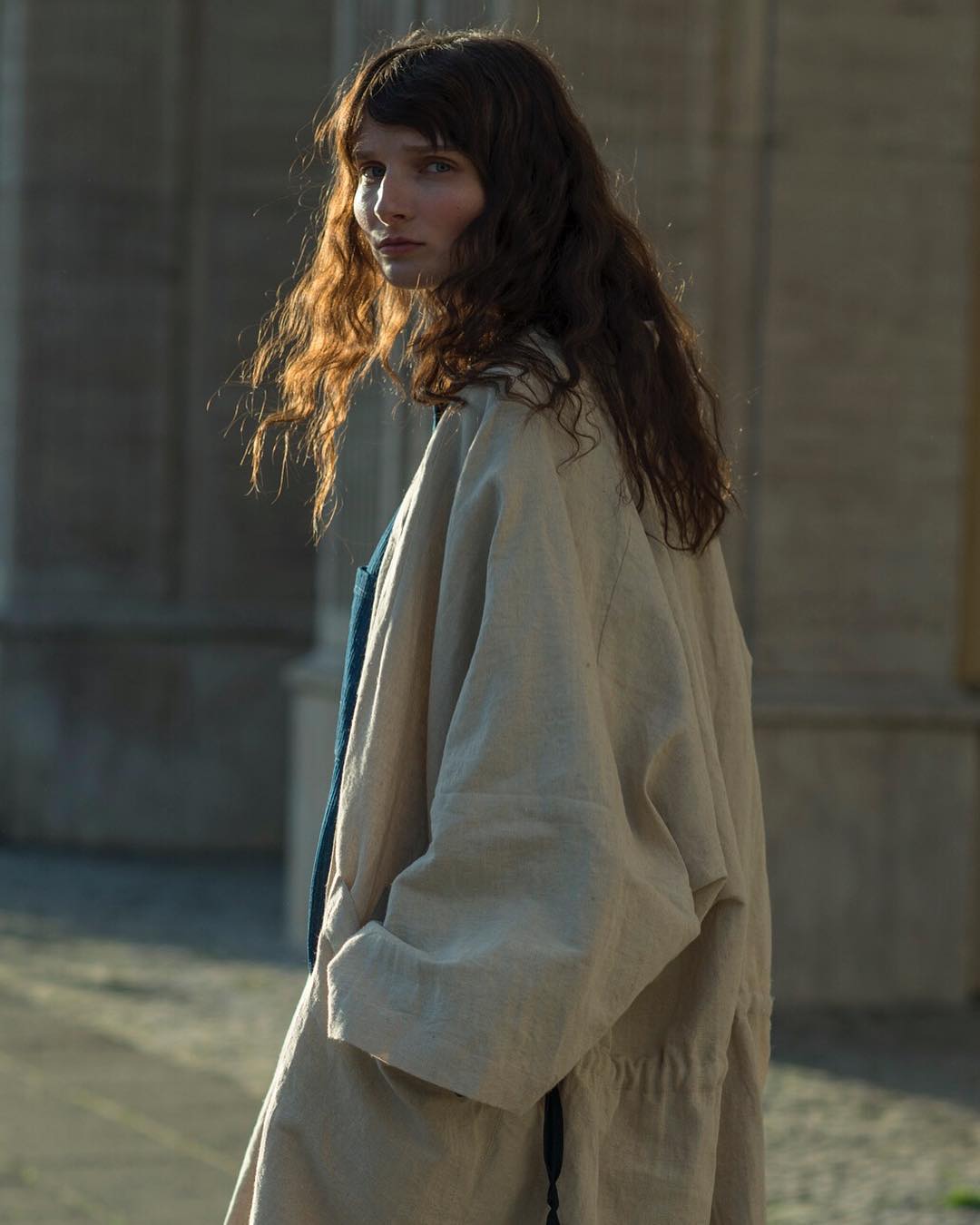 Young woman with brown hair, wearing a beige coat, stands against a stone building in natural sunlight, looking over her shoulder.