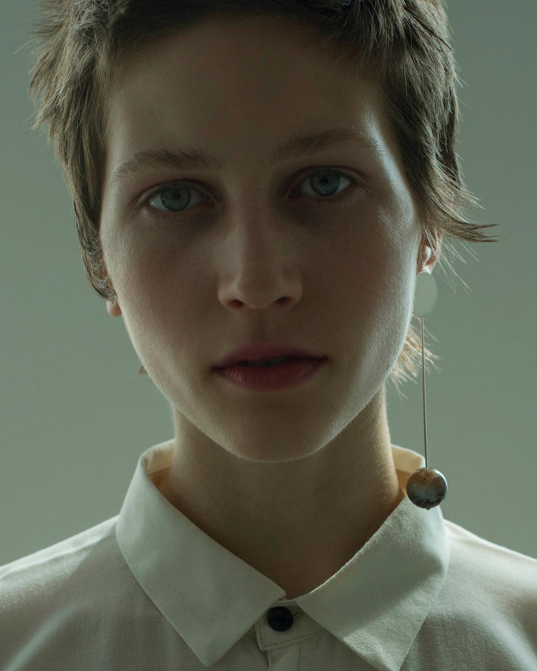 Close-up studio portrait of a young woman with short hair wearing a white button-down shirt and a sculptural sphere earring.