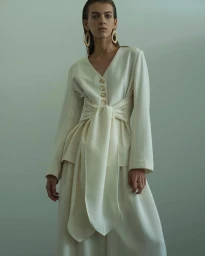Model wearing a cream-colored tied tunic and wide trousers with large geometric earrings against a neutral grey studio wall.