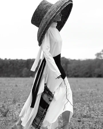 Model wearing an oversized wide-brim woven hat and white dress walking outdoors in a field