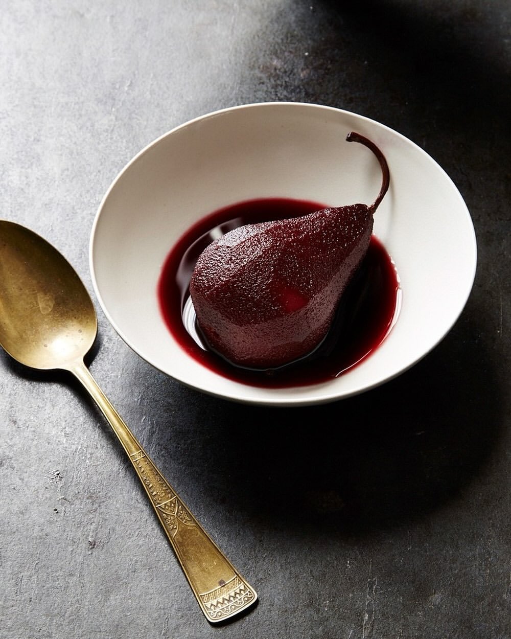 Still life of a poached pear in red wine sauce inside a white bowl next to a golden spoon on a textured dark surface.