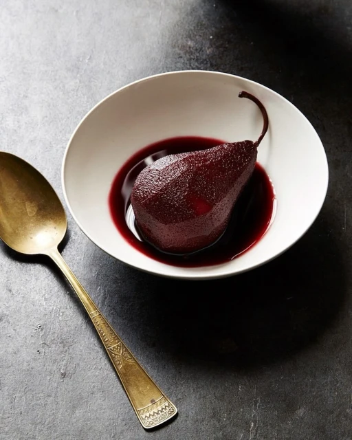 Still life of a poached pear in red wine sauce inside a white bowl next to a golden spoon on a textured dark surface.