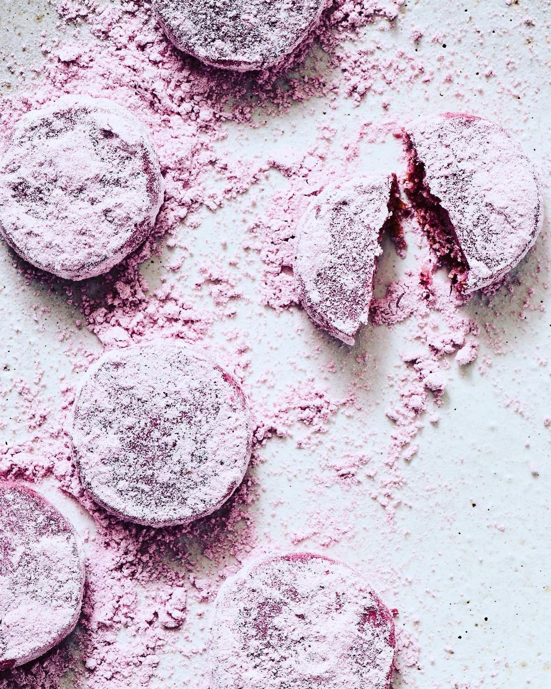 Overhead view of powdered purple cookies, one broken to show inside, with matching purple powder scattered on a textured off-white surface.