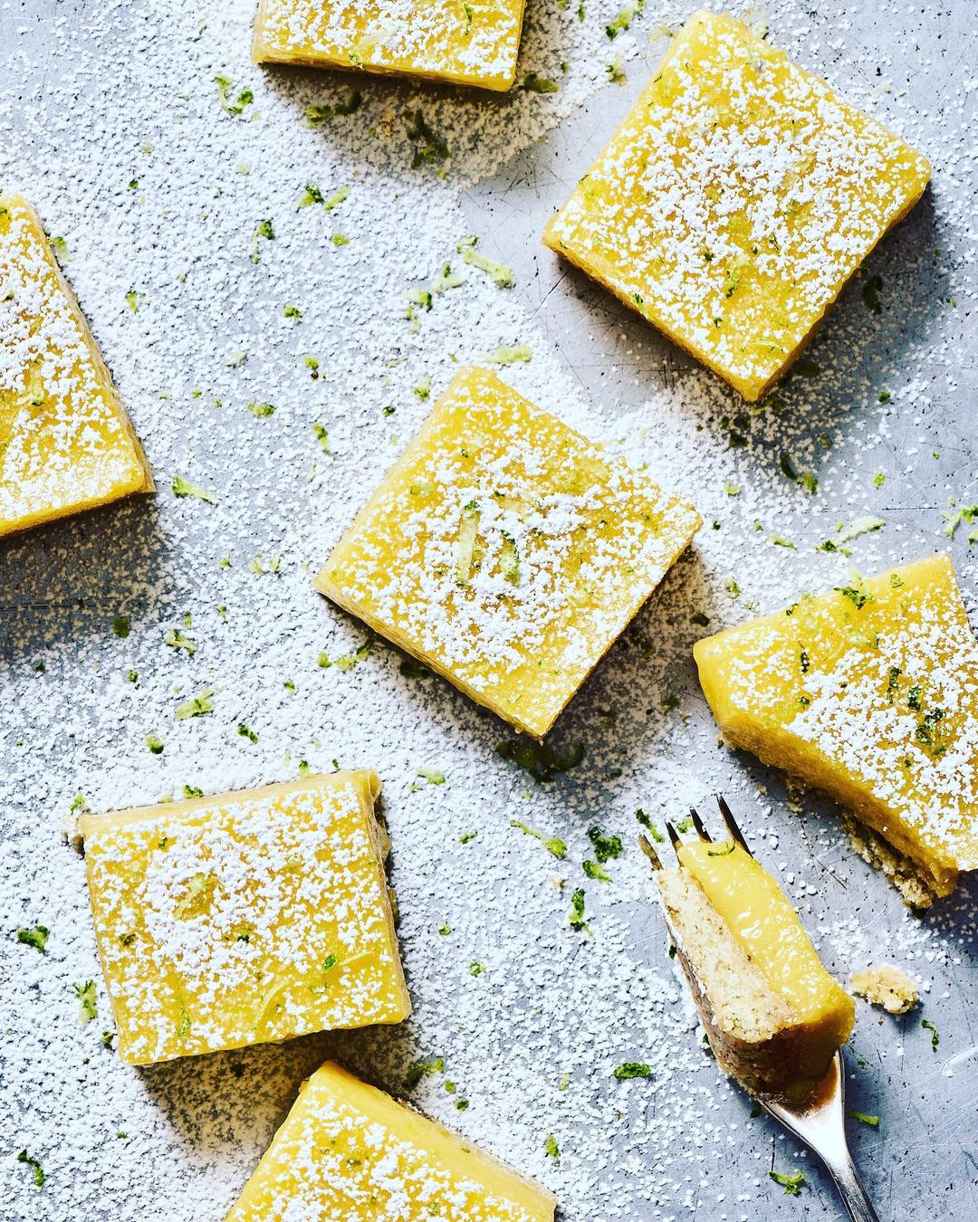 Overhead shot of lemon bars dusted with powdered sugar and lime zest, arranged on a silver surface, with a fork taking a bite.