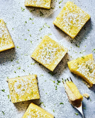 Overhead shot of lemon bars dusted with powdered sugar and lime zest, arranged on a silver surface, with a fork taking a bite.