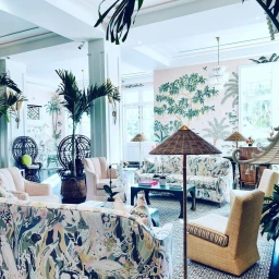 Spacious hotel lounge interior featuring patterned sofas, armchairs, potted palm trees, and tropical wall murals.