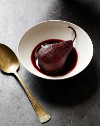 Poached pear in red wine sauce served in a white bowl with a brass spoon on a dark textured surface.