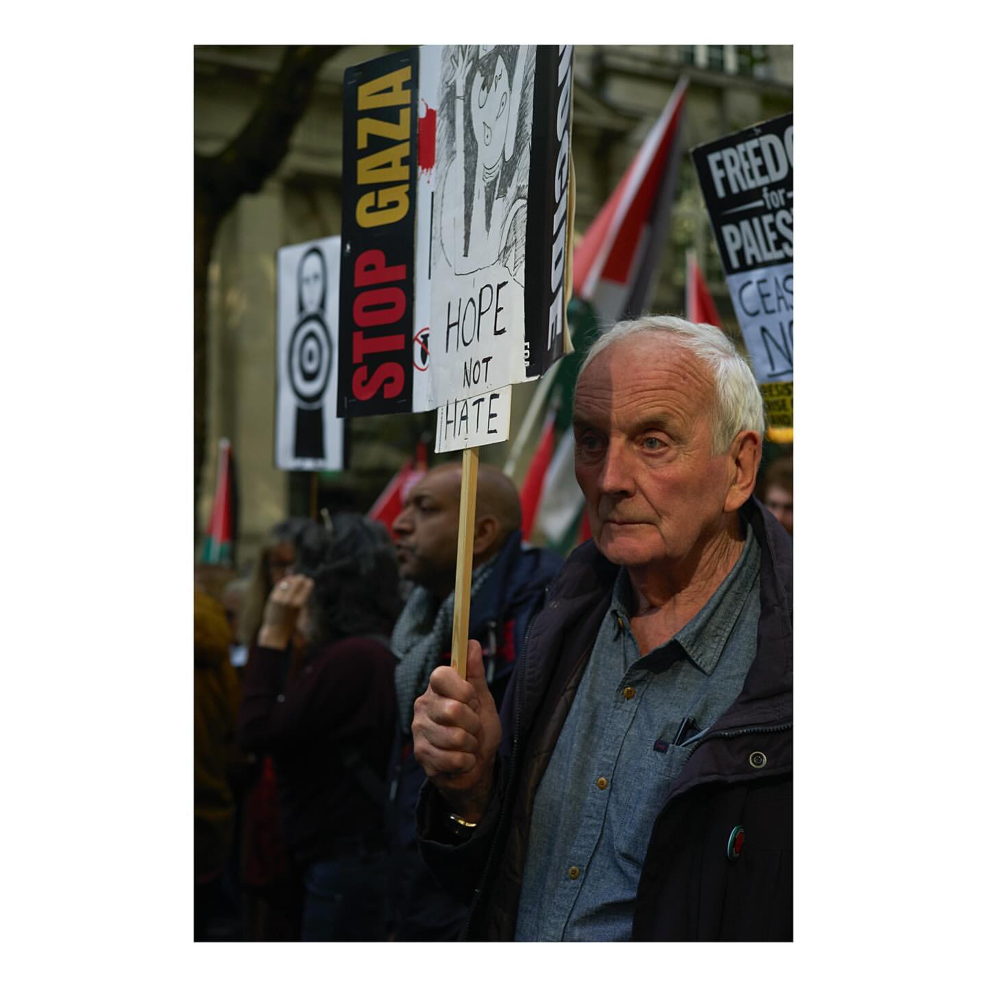 Elderly man holding protest sign 'Hope Not Hate' in a crowd. Demonstration with flags and signs against hate. Urban environment, daylight.