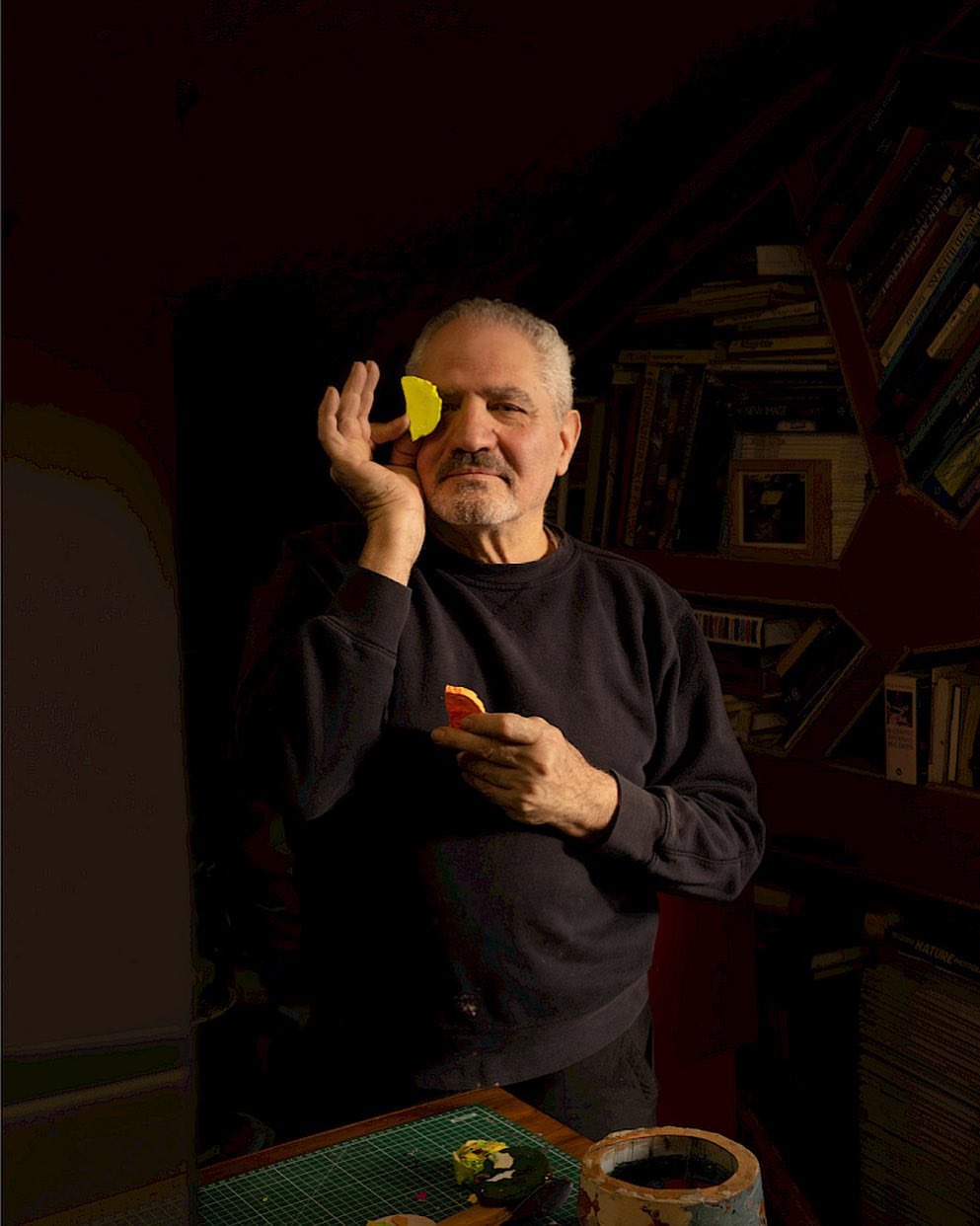 Adult man poses indoors holding yellow and orange discs, covering his eye and standing near a bookcase and pottery.