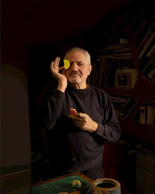 Adult man poses indoors holding yellow and orange discs, covering his eye and standing near a bookcase and pottery.