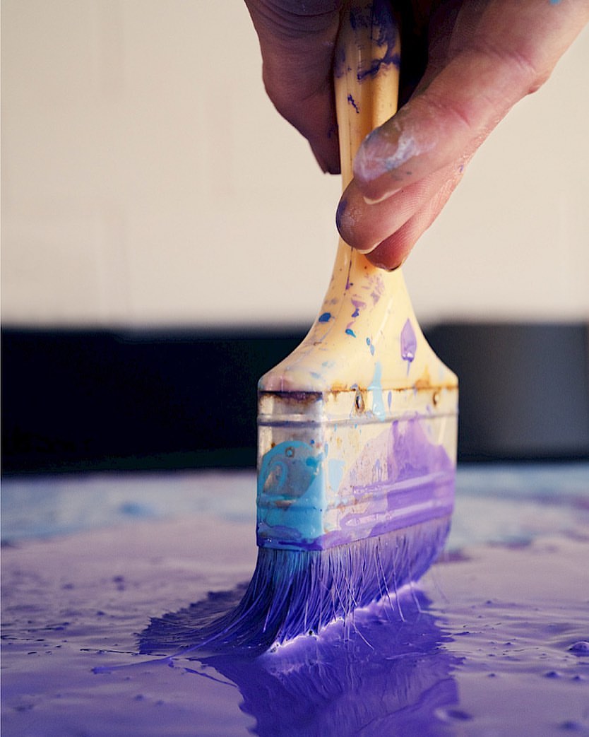 Close-up of a hand holding a paintbrush loaded with purple paint, poised over a surface covered in wet purple paint.